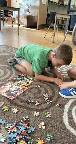 2 JKK children sitting on the carpet with completed puzzles and puzzle pieces surrounding them. One child is crouched forward, the other sitting on their knees