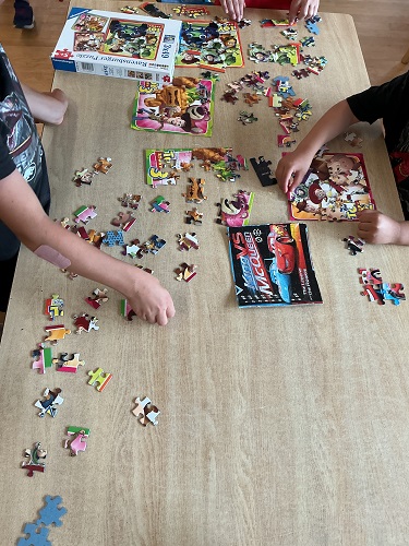 3 school age children standing around a table with posters of what the puzzles will look like with multiple puzzle pieces around the table