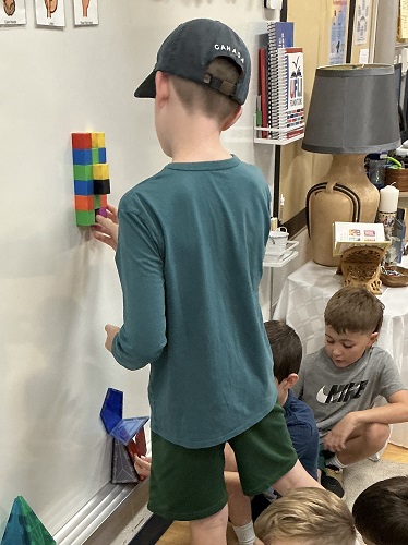 5 School age children around the white board with magnets, one child standing at the board, the rest are crouched around.