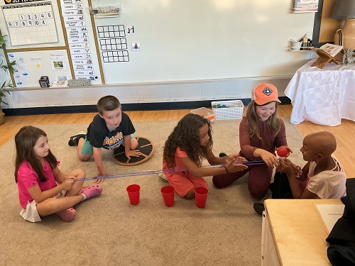 5 School Age children sitting on the carpet, one child on his knees looking at the camera, another child sitting cross legged holding the string, another child sitting cross legged holding the opposite side of the string, and 2 children balancing the ball on the string.