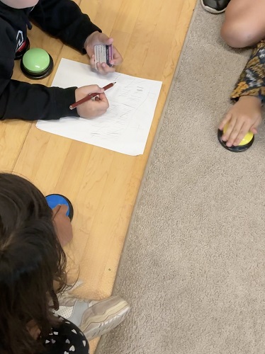 3 School Age children sitting around each other, one child has a paper in front of them, holding a pencil in one hand and a card in the other, each child has coloured buzzer in front of them