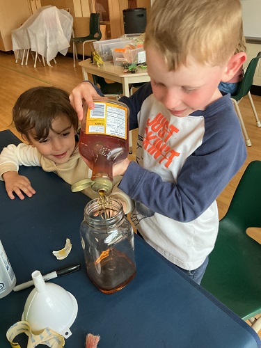 Child pouring corn syrup into a glass jar while another child watches from behind