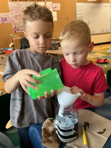 Child pouring water into a funnel into a glass jar while another child holds the funnel