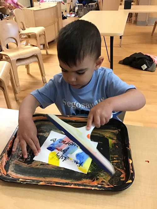 Preschool boy sitting at table with a squeegee spreading blue and yellow paint on paper